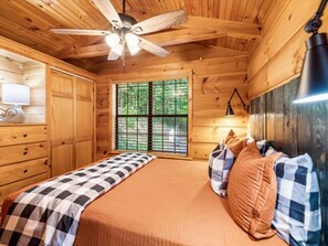 Une chambre dans une cabane en bois, avec un lit, un ventilateur de plafond, une fenêtre et une lampe.