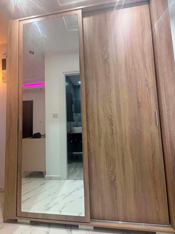 Un armario de madera con puerta espejada, suelo de baldosas y un baño a la vista en el fondo.