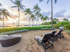 Un espace de détente aménagé au bord de la plage, avec des chaises longues et un coin salon abrité, entouré de palmiers et offrant une vue sur l’océan.