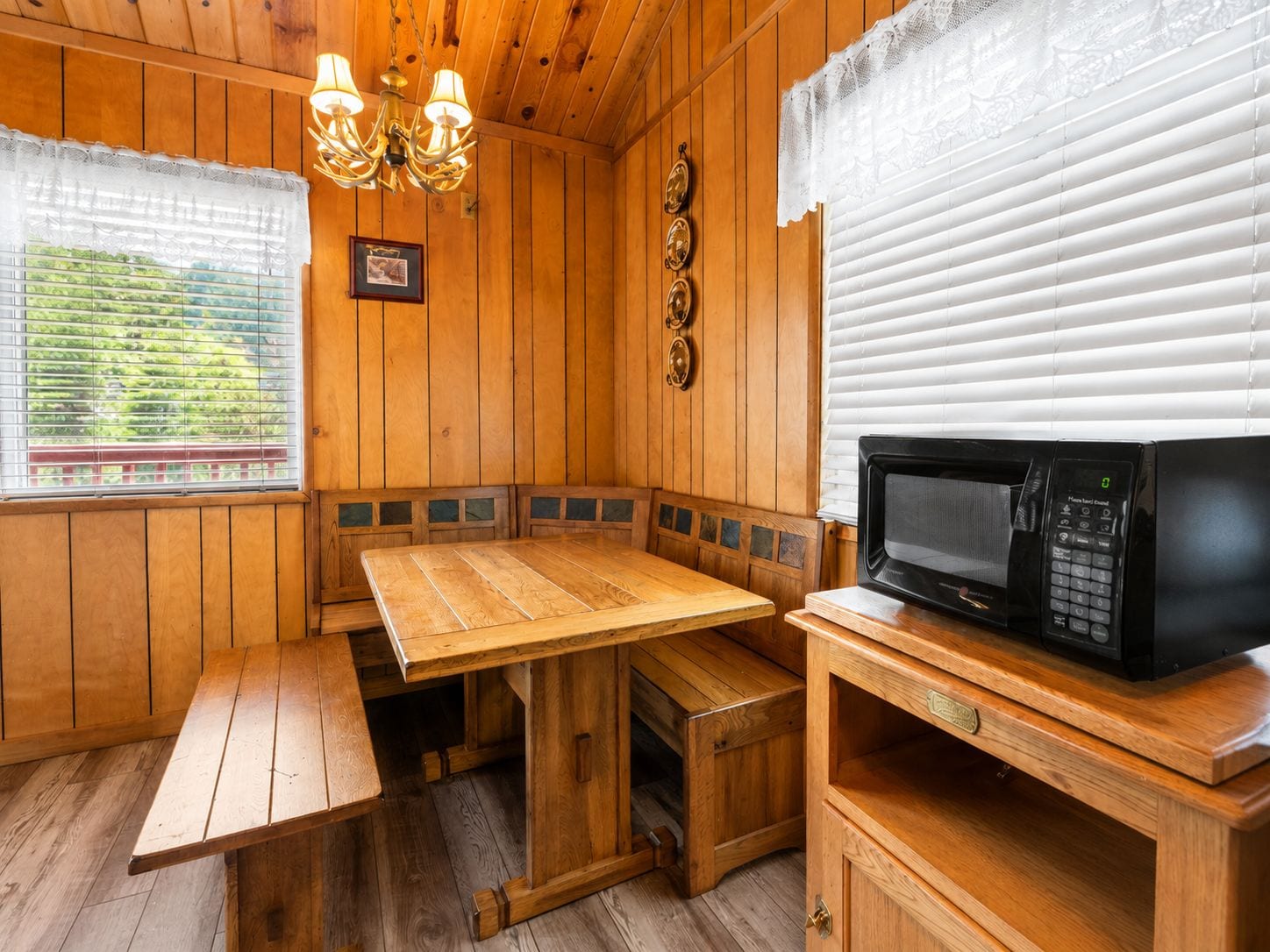 Interior de una cabaña de madera con una mesa de madera, bancos, una araña, un microondas y una ventana con persianas.