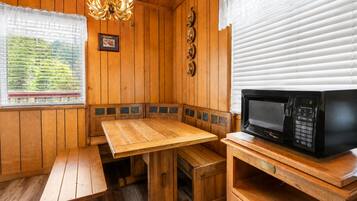 Interior de una cabaña de madera con una mesa de madera, bancos, una araña, un microondas y una ventana con persianas.