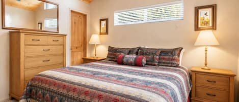 A bedroom with a wooden ceiling, a large bed with patterned bedding, a dresser, two bedside tables with lamps, a window with blinds, and a framed picture on the wall.