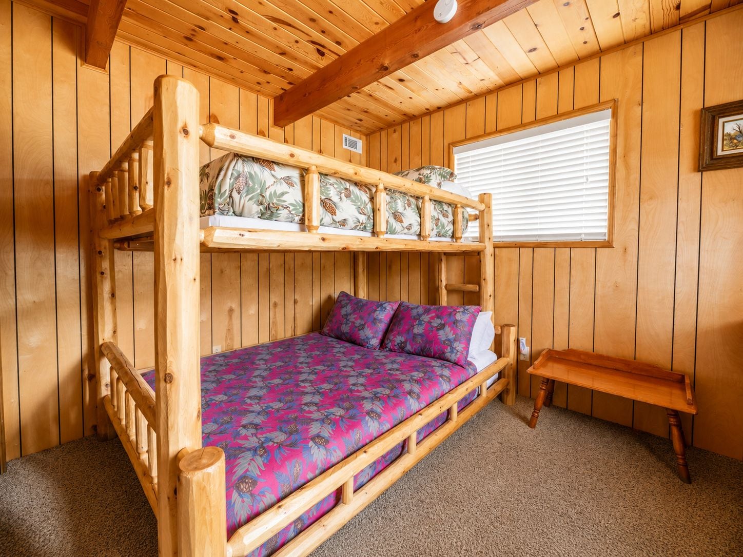A bunk bed with a floral bedspread and pillows, a wooden bench, and a window with blinds.