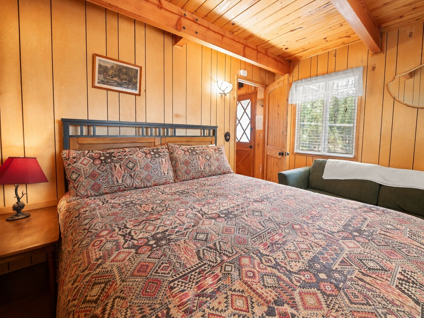Una habitación de madera con una cama, una mesita de noche, una lámpara, una ventana con persianas y una puerta.
