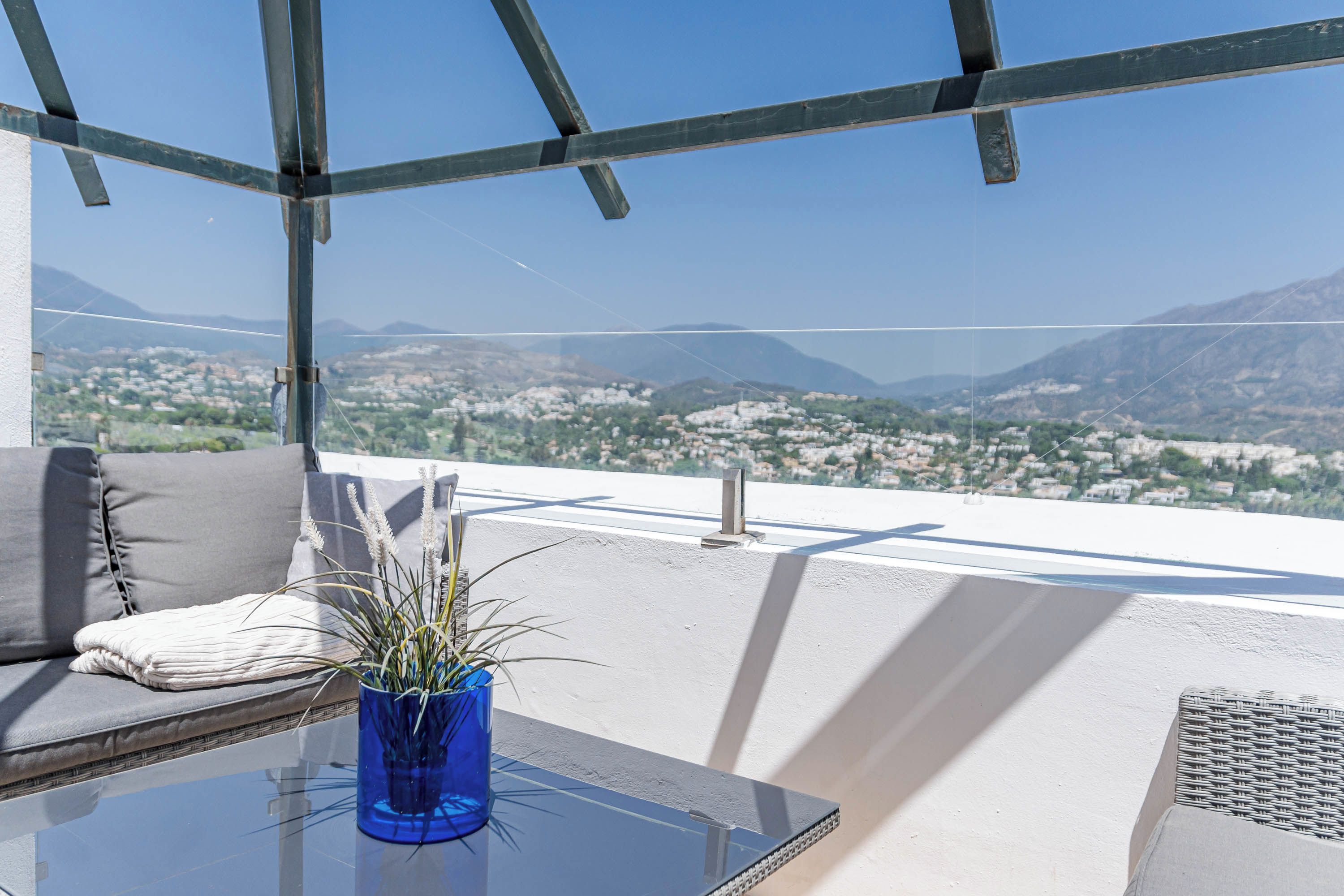 A balcony with a glass table, a blue vase with plants, a cushion, and a view of a cityscape with mountains.