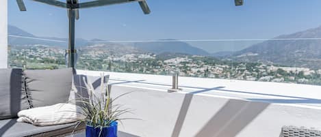 Un balcon avec une table en verre, un vase bleu contenant des plantes, un coussin et une vue sur un paysage urbain avec des montagnes.