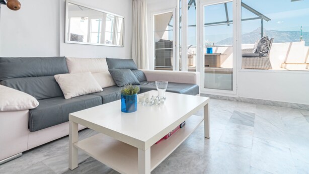 A modern living room with a grey sofa, white coffee table, and a view of the outdoors through large windows. - Sam65i Central 2 Bed Penthouse in Nueva Andalucia (Marbella)