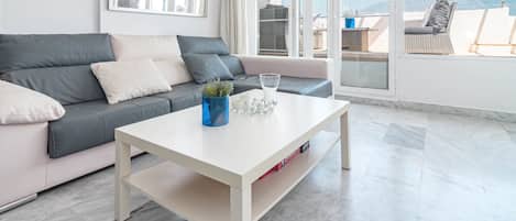 A modern living room with a grey sofa, white coffee table, and a view of the outdoors through large windows.