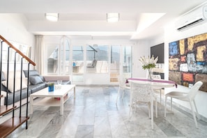 A modern living room with a sofa, coffee table, and chairs, a dining area with a table and chairs, and a large window with a view of the outdoors. - Sam65i Central 2 Bed Penthouse in Nueva Andalucia (Marbella)