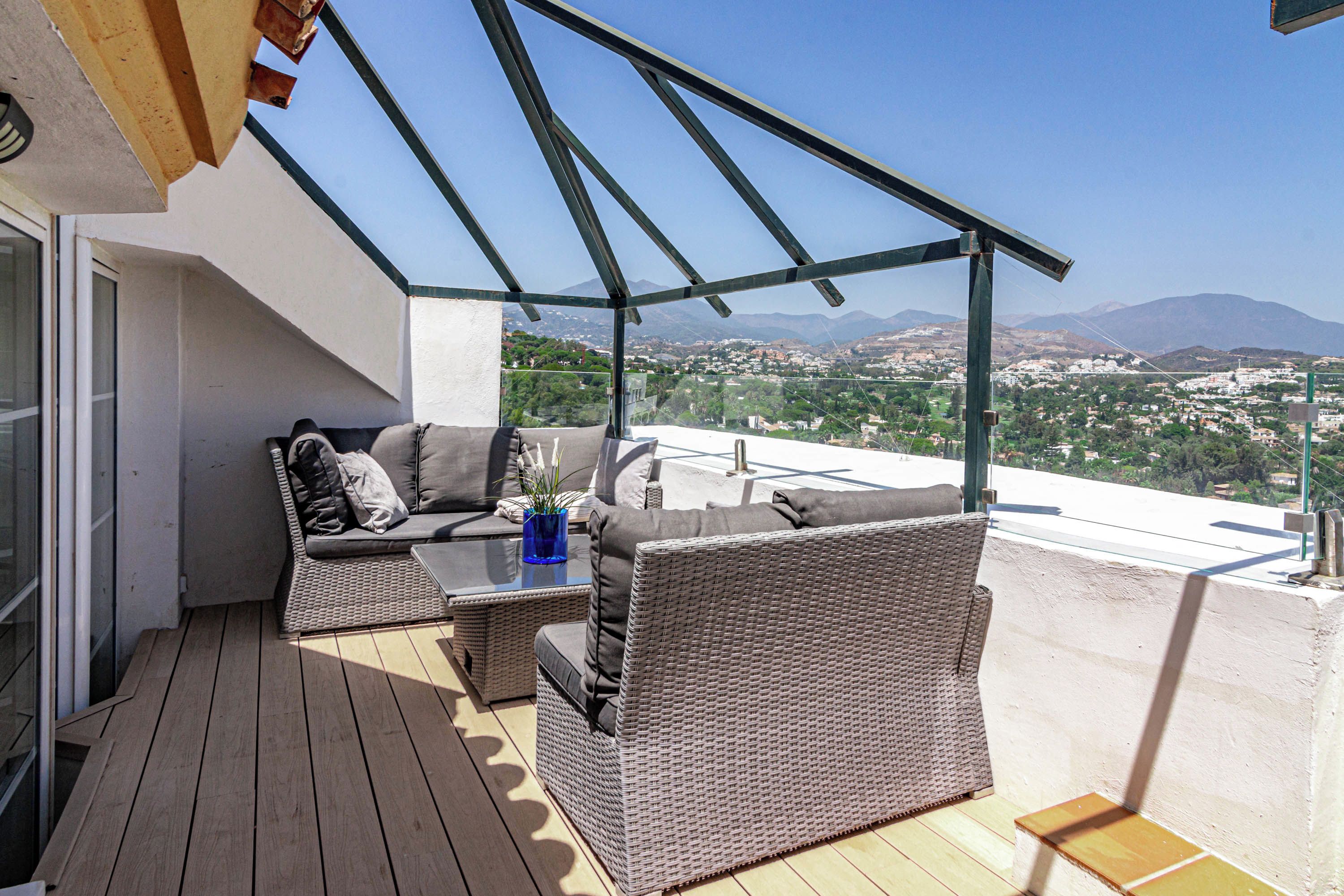 A balcony with wicker furniture, a glass table, and a view of a cityscape and mountains.