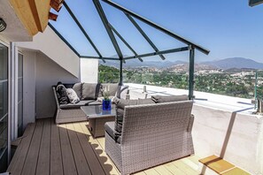 Un balcón con mobiliario de mimbre, una mesa de vidrio y vistas a un paisaje urbano y montañas.