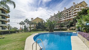 A swimming pool with a metal ladder, surrounded by a well-maintained lawn and palm trees. - Sam65i Central 2 Bed Penthouse in Nueva Andalucia (Marbella)