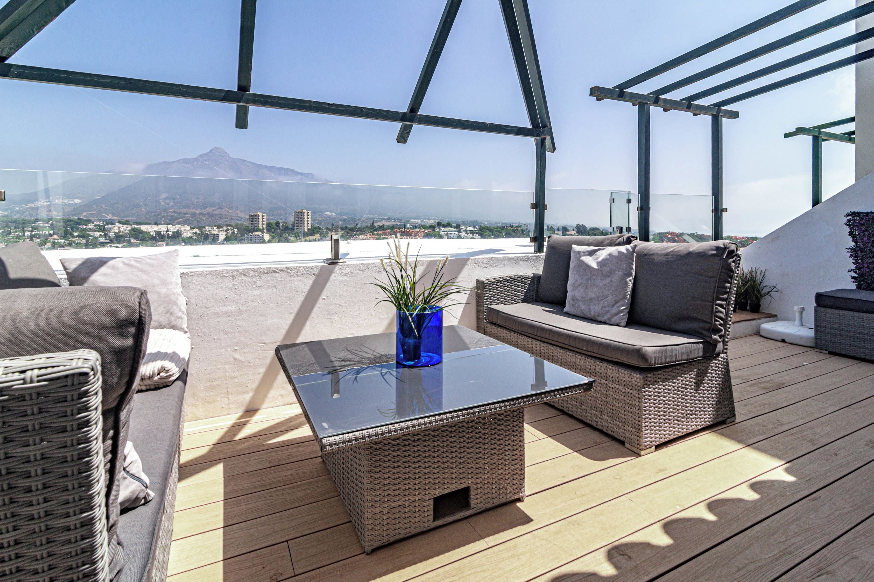 A rooftop terrace with wicker furniture, a glass coffee table, and a view of a mountain.