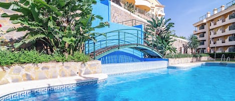 Una piscina con un muro de piedra y una estructura azul, rodeada de vegetación y edificios.