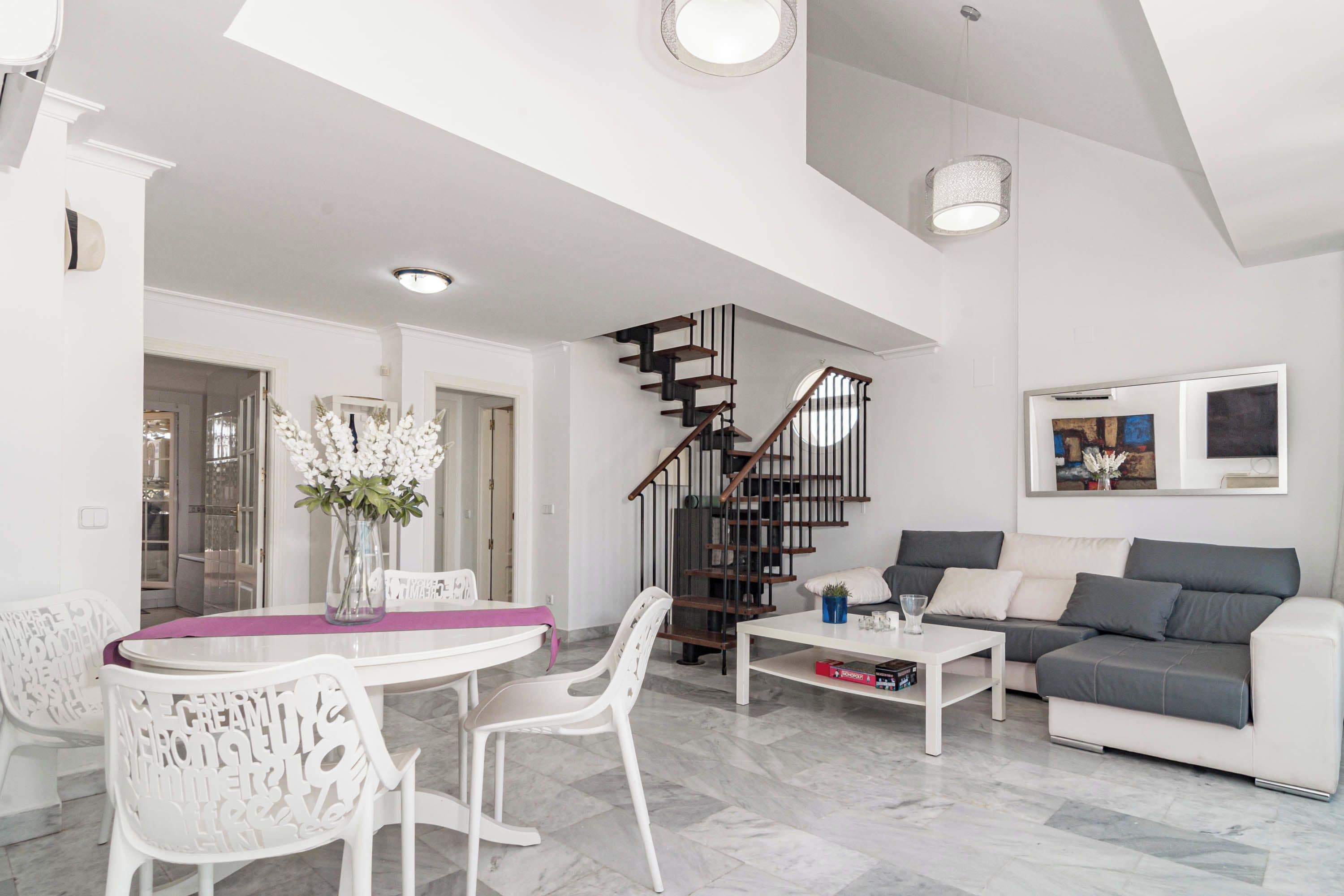 A modern living room with a white sofa, a dining table with chairs, a staircase, and a chandelier.