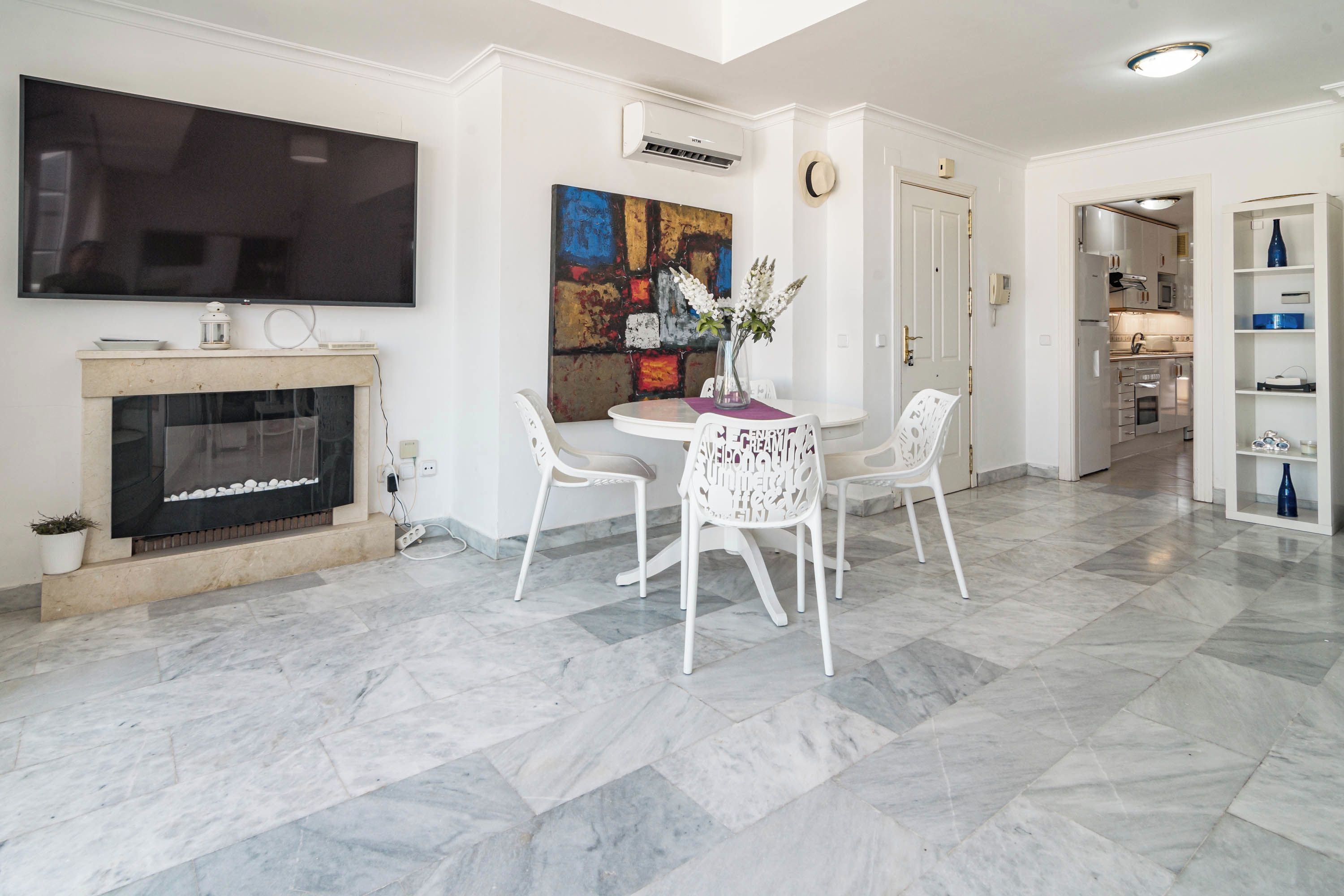 Una sala moderna con televisor de pantalla plana, chimenea, comedor con sillas blancas y cocina visible a través de una puerta abierta.