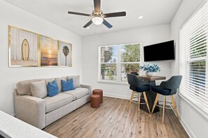 A modern living room with a sofa, coffee table, and chairs, a ceiling fan, and a large window with blinds.