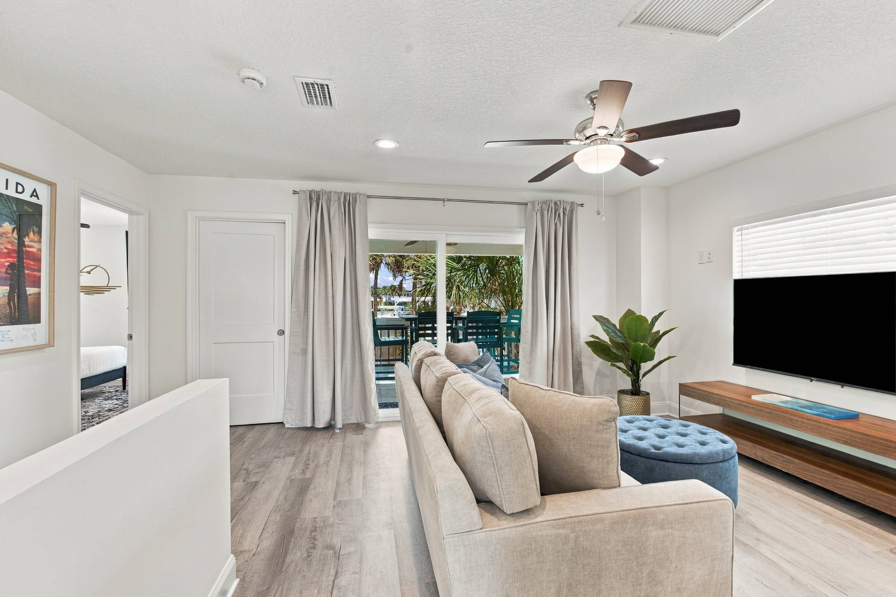 A modern living room with a sofa, a ceiling fan, a TV, and a view through a glass door.