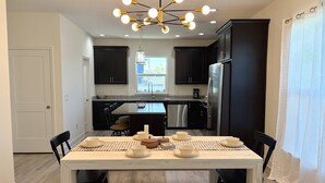 A modern kitchen with dark cabinetry, a stainless steel refrigerator, and a dining area with a wooden table set for four. - Private Beach Access, Beautiful Ocean Views, Balcony, Brand New Furnishings, $300 in Free Tickets! (St. Augustine)