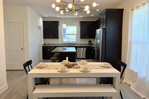 A modern kitchen with dark cabinetry, a stainless steel refrigerator, and a dining area with a wooden table set for four. - Private Beach Access, Beautiful Ocean Views, Balcony, Brand New Furnishings, $300 in Free Tickets! (St. Augustine)