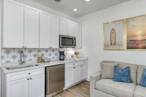 A modern kitchen with white cabinets, a stainless steel dishwasher, and a microwave. There is a coffee maker and a surfboard-themed wall art.
