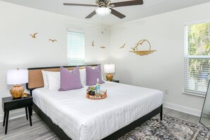 A bedroom with a bed, two nightstands, a ceiling fan, a window with blinds, and wall art. - Private Beach Access, Ocean Views, Hot Tub, Balcony, Brand New Furnishings, $300 in Free Tickets! (St. Augustine)