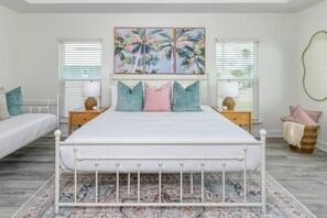 A bedroom with a large bed, two bedside tables, a decorative rug, and a wall with a tropical painting.