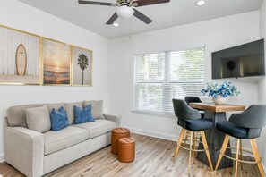 A modern living room with a sofa, ottomans, a dining area with chairs, a ceiling fan, and a TV mounted on the wall.