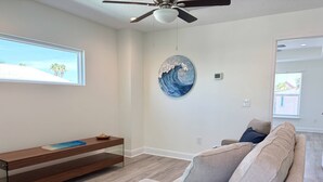 A modern living room with a ceiling fan, a large window with a view of palm trees, a round wall art piece, a wooden bench, and a grey sofa with cushions. - Private Beach Access, Beautiful Ocean Views, Balcony, Brand New Furnishings, $300 in Free Tickets! (St. Augustine)
