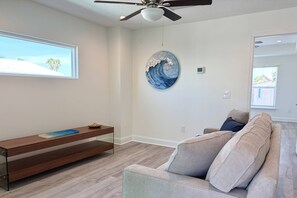 A modern living room with a ceiling fan, a large window with a view of palm trees, a round wall art piece, a wooden bench, and a grey sofa with cushions. - Private Beach Access, Beautiful Ocean Views, Balcony, Brand New Furnishings, $300 in Free Tickets! (St. Augustine)