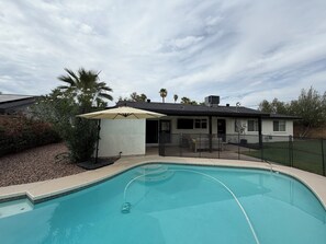 A swimming pool with a patio area, a covered seating area, and a house in the background. - The Cove: Poolside Getaway near PV & Camelback (Phoenix)