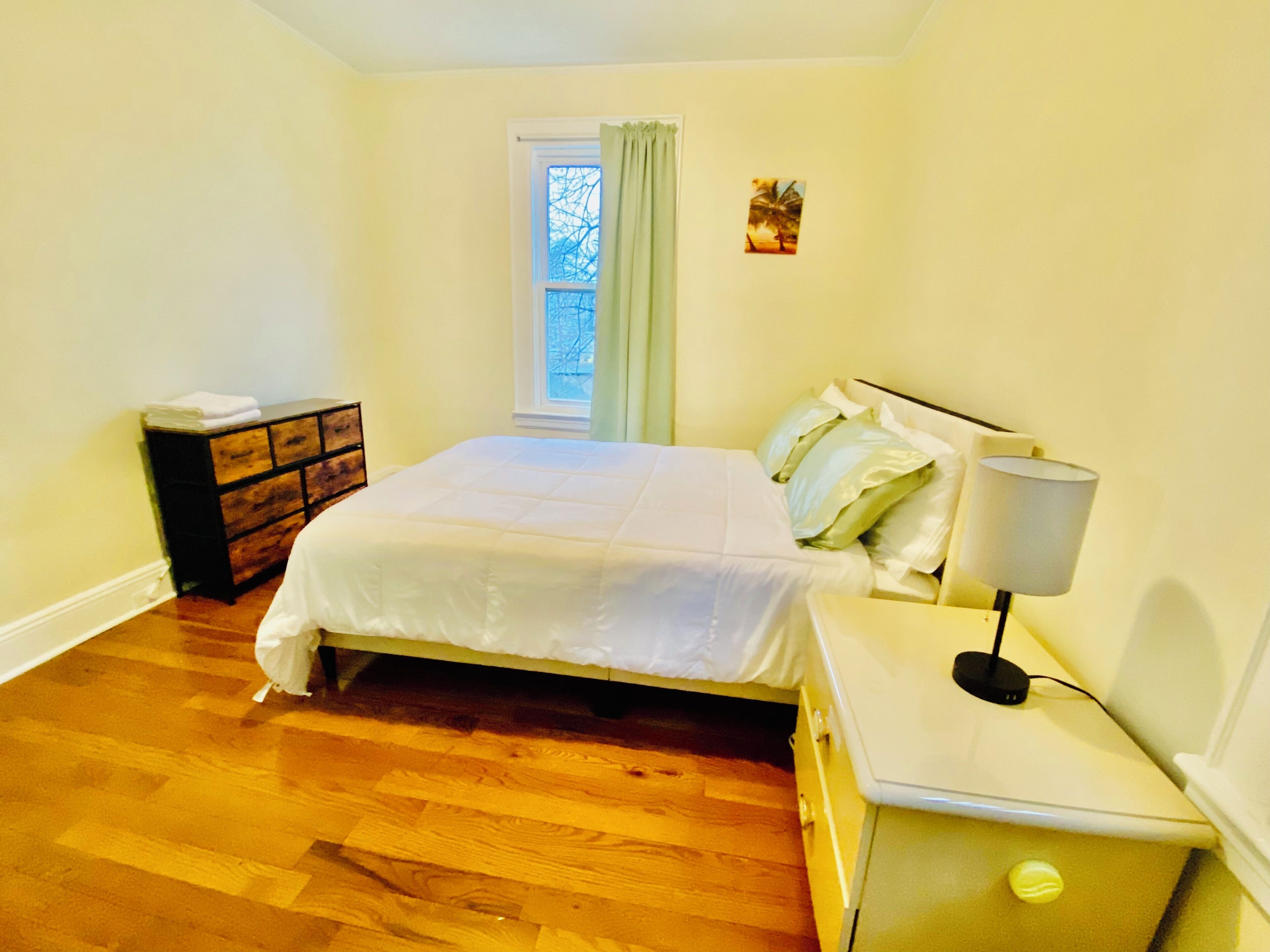 Ein Schlafzimmer mit Bett, Nachttisch, Lampe, Kommode und einem Fenster mit Vorhängen.