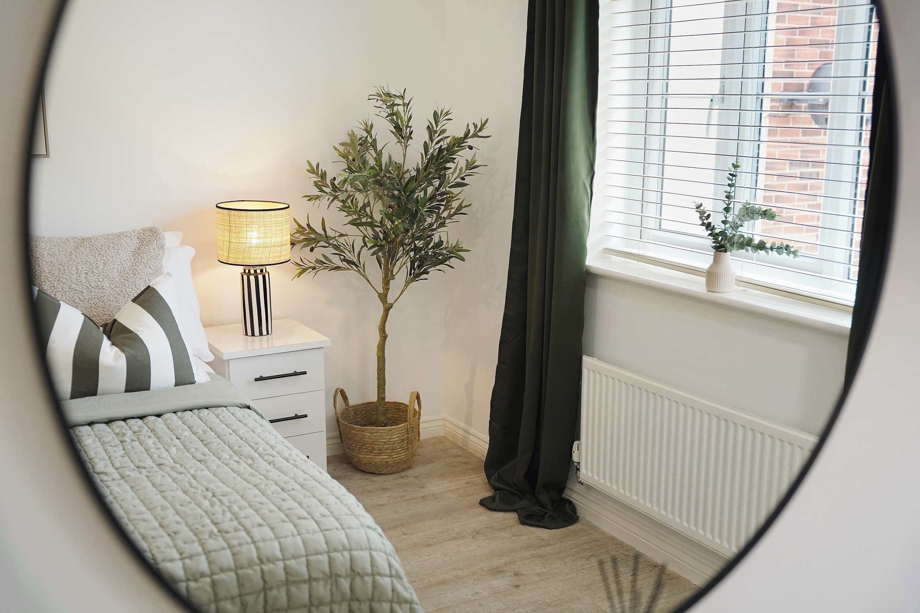 A bedroom with a bed, a bedside table with a lamp, a potted plant, a window with blinds, and a radiator.