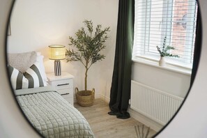 A bedroom with a bed, a bedside table with a lamp, a potted plant, a window with blinds, and a radiator. - Modern 4BR Stay Driveway Parking By Stay With Us (Middlesbrough)
