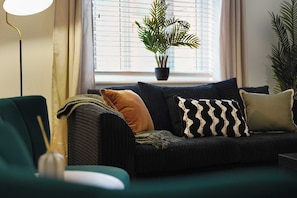 Ein modernes Wohnzimmer mit einem dunkelgrünen Sofa, mehreren Kissen, einer Topfpflanze am Fensterbrett und einer Stehlampe.