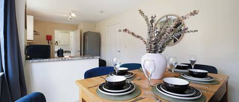 A dining area with a wooden table set for four, a vase with dried flowers, and a kitchen in the background.