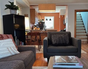 Ein Wohnzimmer mit einer Couch, einem Couchtisch mit Büchern, einer Küche im Hintergrund und einer Treppe.