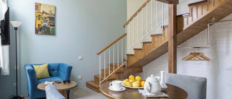 A modern living room with a blue sofa, a round wooden table with a white cup and fruit, and a staircase with wooden railings.
