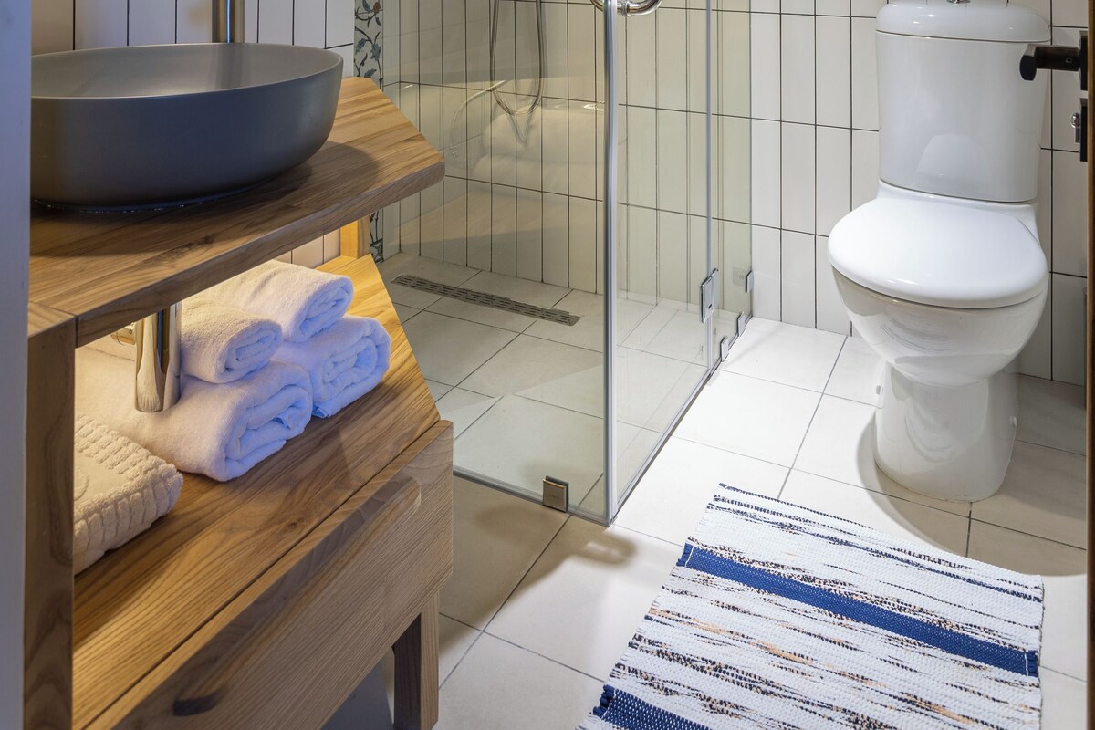 A modern bathroom with a glass-enclosed shower, a round mirror, a wooden vanity, and a white toilet.