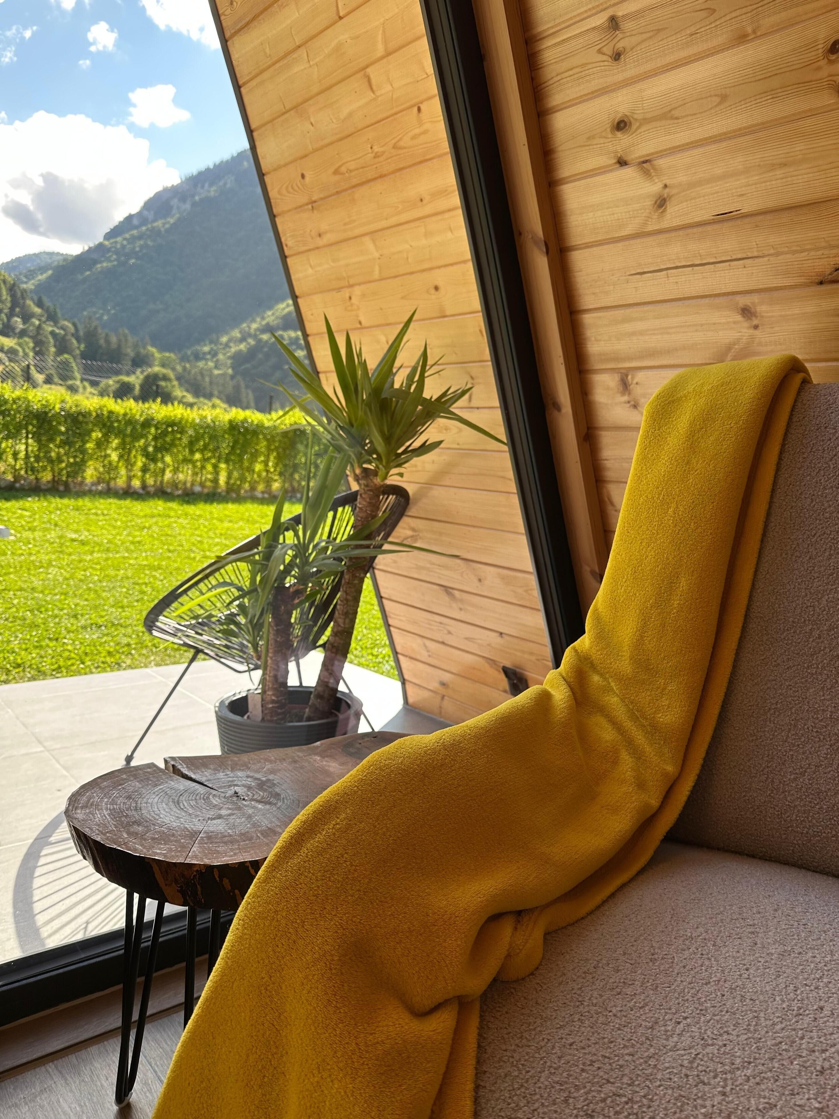 Une pièce avec vue sur un paysage verdoyant, une plante en pot et un plaid jaune sur un canapé.