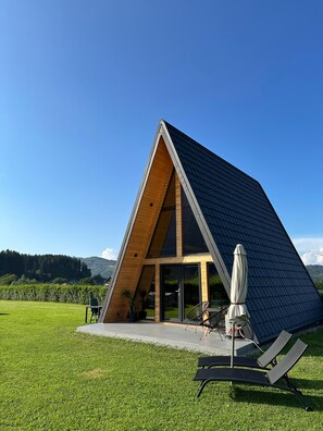 Ein modernes, dreieckiges Gebäude mit geneigtem Dach, großen Glasüren und einer Terrasse mit Gartenmöbeln.