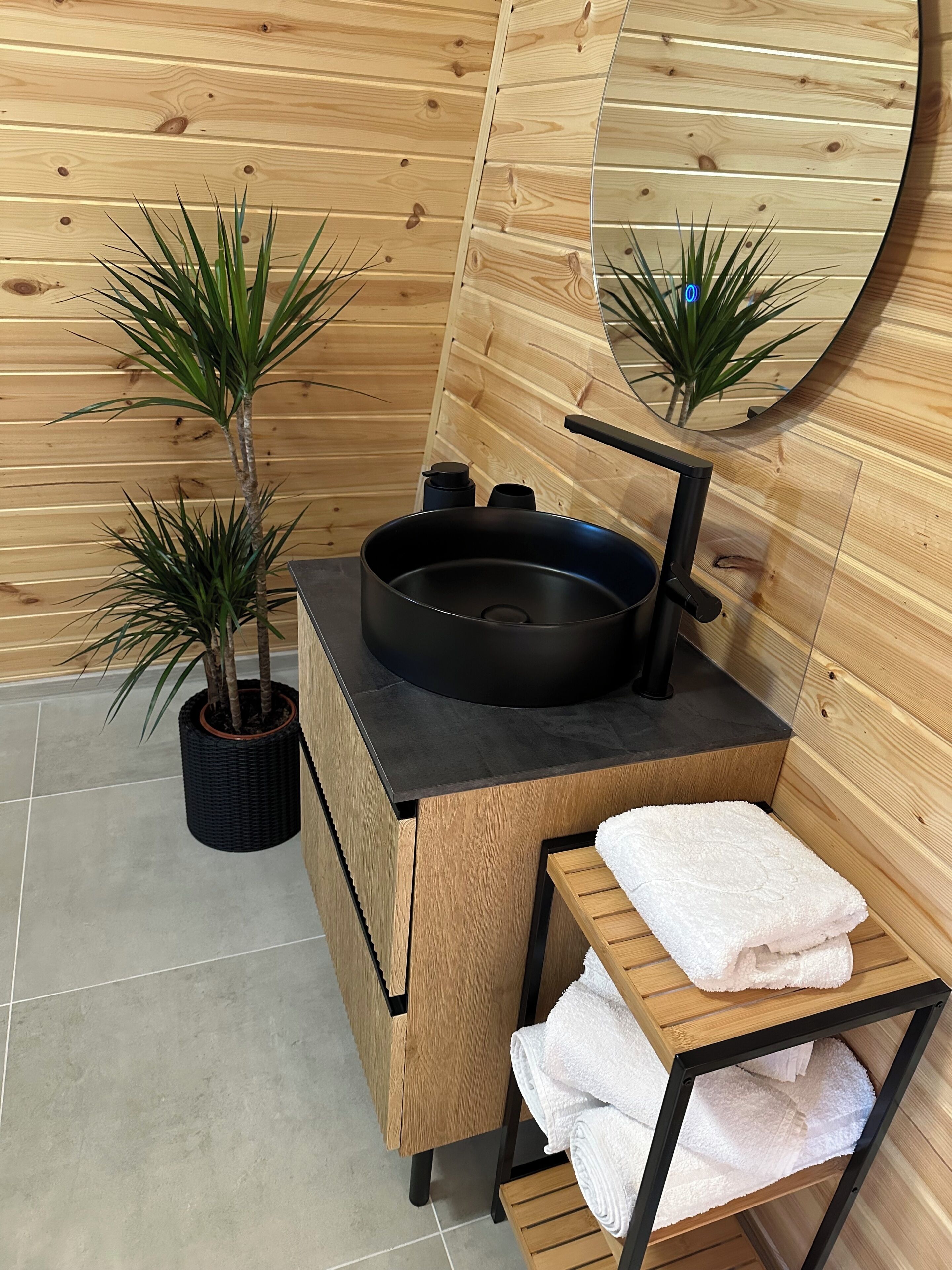 Ein Badezimmer mit einem Holzbadezimmermöbel, einem schwarzen Waschbecken und einem Spiegel. Zwei Blumentöpfe und ein Handtuchhalter mit gefalteten Handtüchern sind vorhanden.