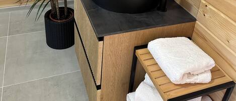 Une salle de bain avec un meuble-lavabo en bois, un évier noir et un miroir. On y trouve deux plantes en pot et un porte-serviettes avec des serviettes pliées.