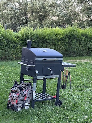 Un barbecue portable doté d’une table d’appoint, posé sur l’herbe, avec un sac de charbon de bois et une brosse à proximité.