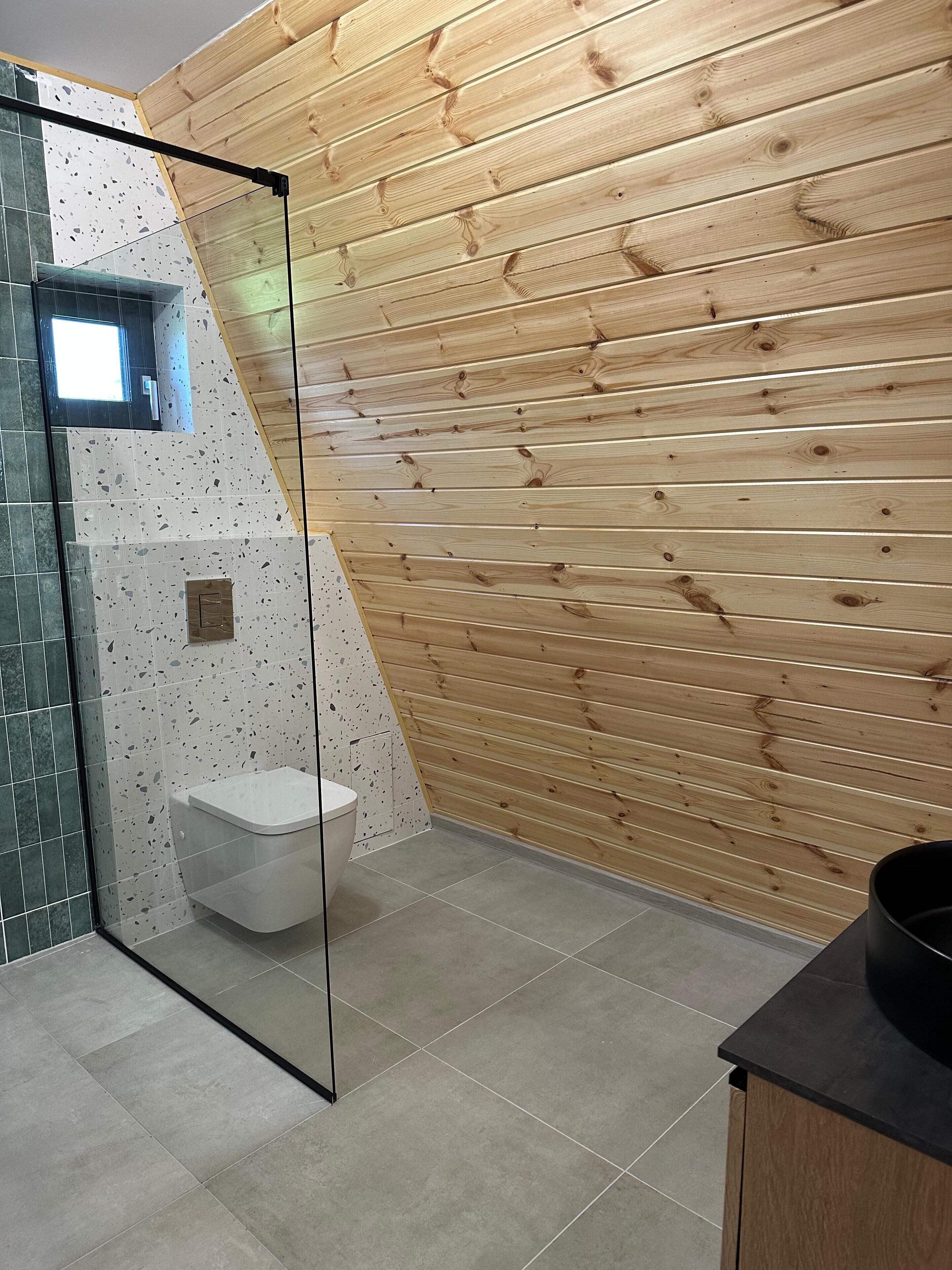 Une salle de bain avec un mur en lambris de bois, une douche en verre et des toilettes blanches.
