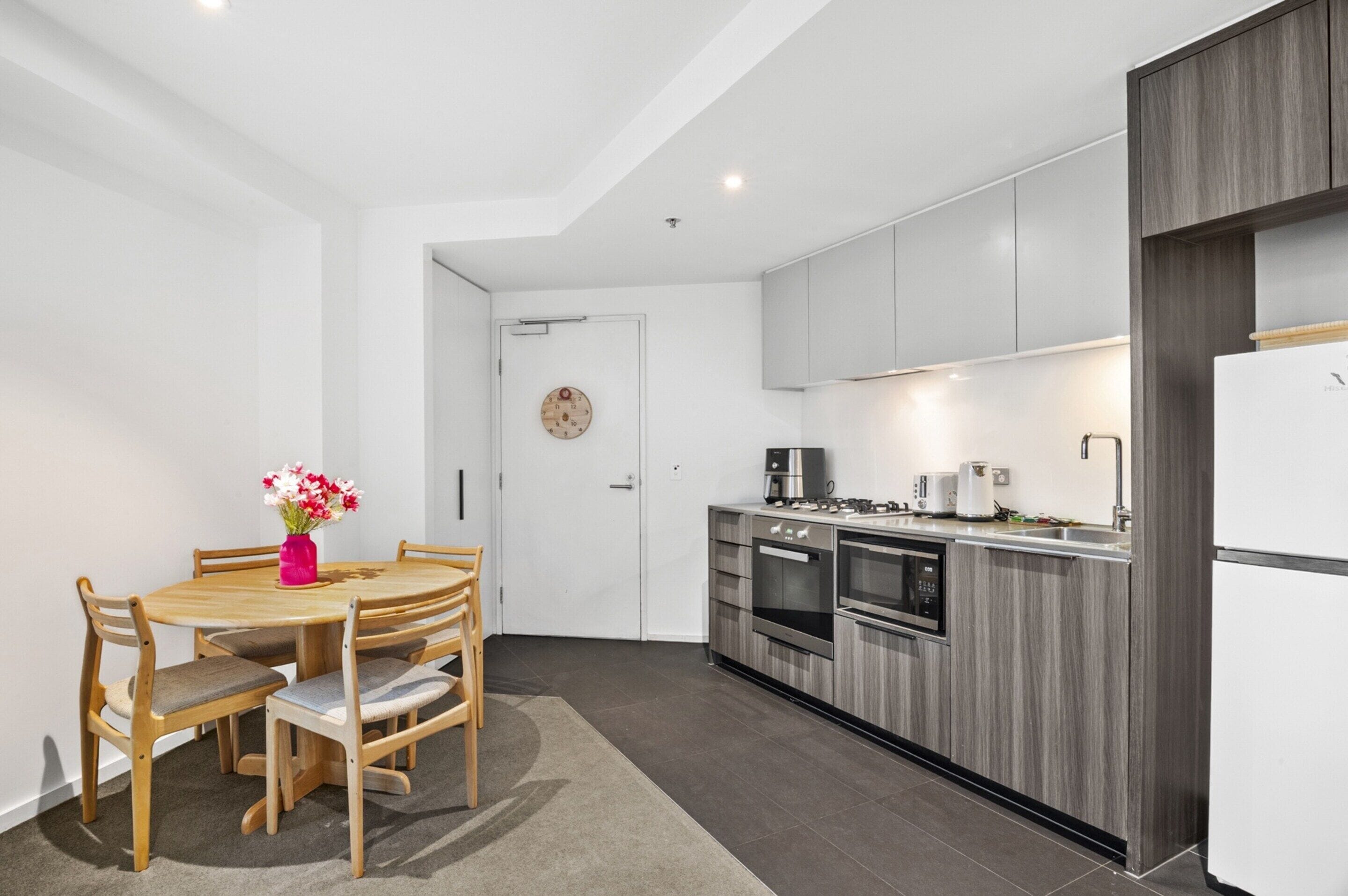 Une cuisine compacte dotée d’appareils électroménagers modernes, un coin repas avec une table et des chaises en bois, et une petite horloge au mur.