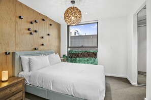 Ein modernes Schlafzimmer mit grauem Polsterbett, einer Holzwand mit Nummernblöcken, einer hängenden, kugelförmigen Leuchte und Blick auf ein Gebäude durch ein Fenster mit Jalousien.