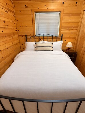 A single bed with a metal frame, a window with blinds, and a small bedside table with a lamp. - Eagles Nest Lodge (Butternut)