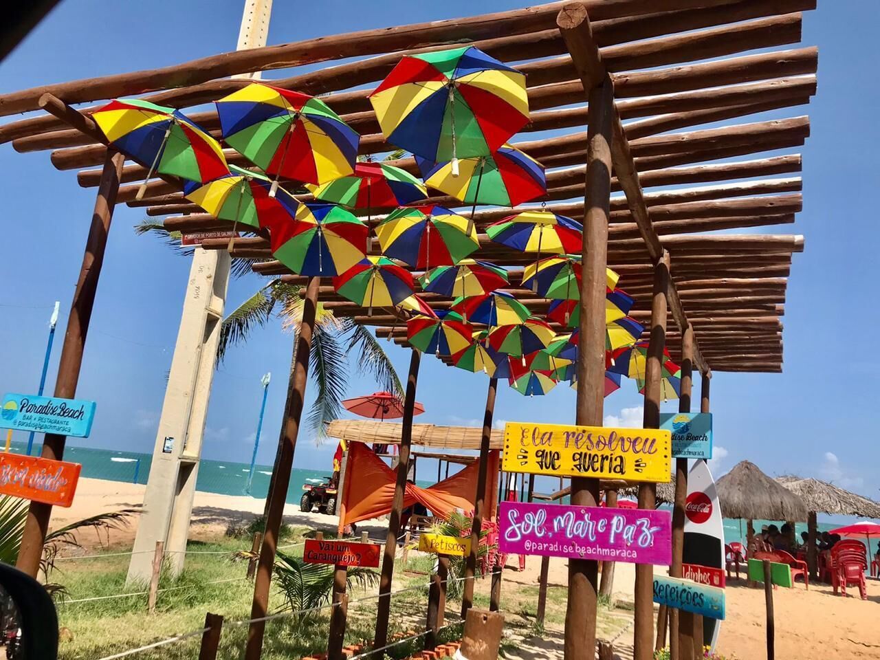 Une zone de plage agrémentée de nombreux parasols colorés, de structures en bois et de panneaux indiquant les activités proposées sur la plage.