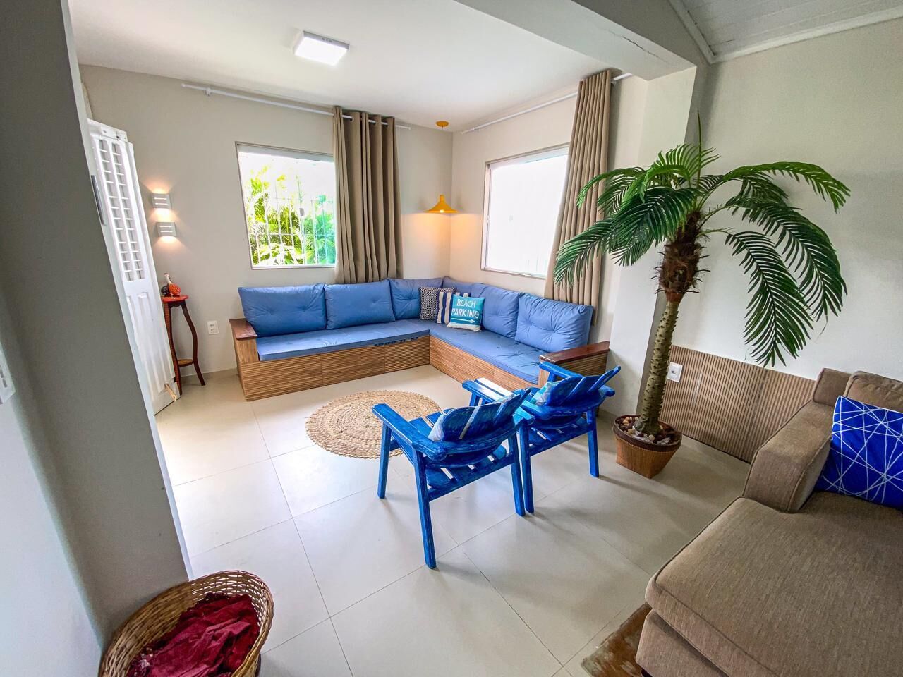 A living room with a sectional sofa, a coffee table, and a blue armchair. There is a potted palm tree and a window with curtains.